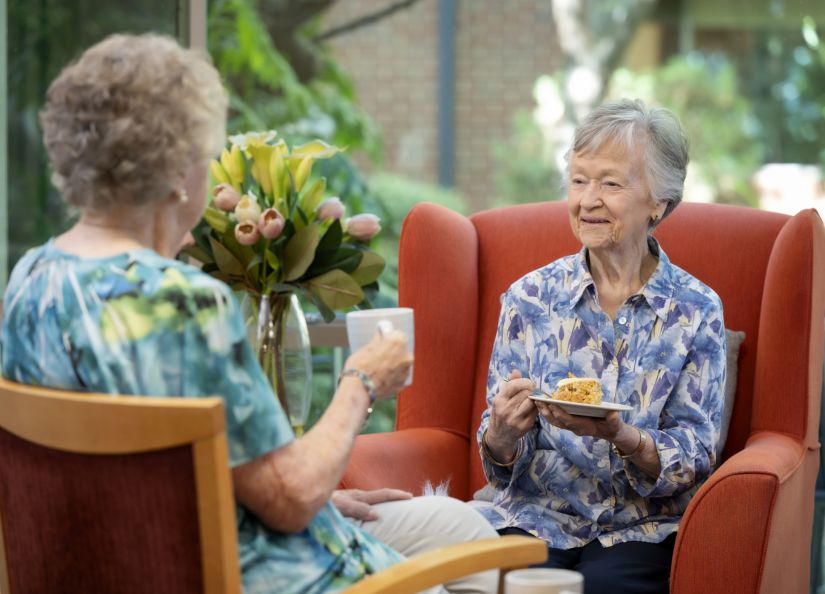 Two residents seated in chairs, enjoying a meal together and smiling.